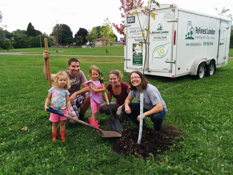 Small group planting a tree in a park, with ReForest London trailer in the background.