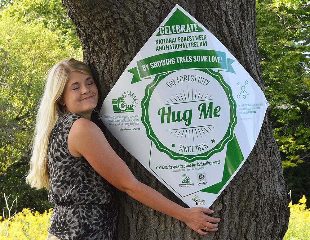 Woman hugging a tree with a sign saying, "Hug Me."