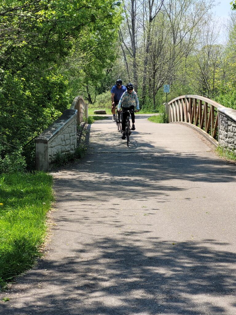 Two cyclists cross a small bridge over a small stream on the Thames Valley Parkway.