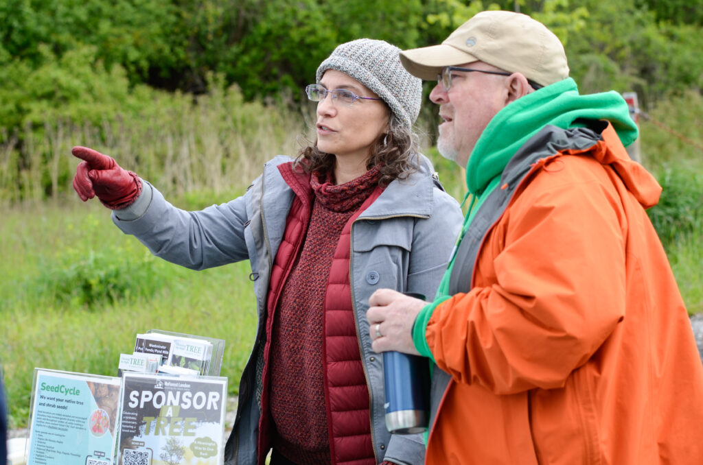 Volunteer from ReForest London instructs a man visiting an information table set up near a forest.
