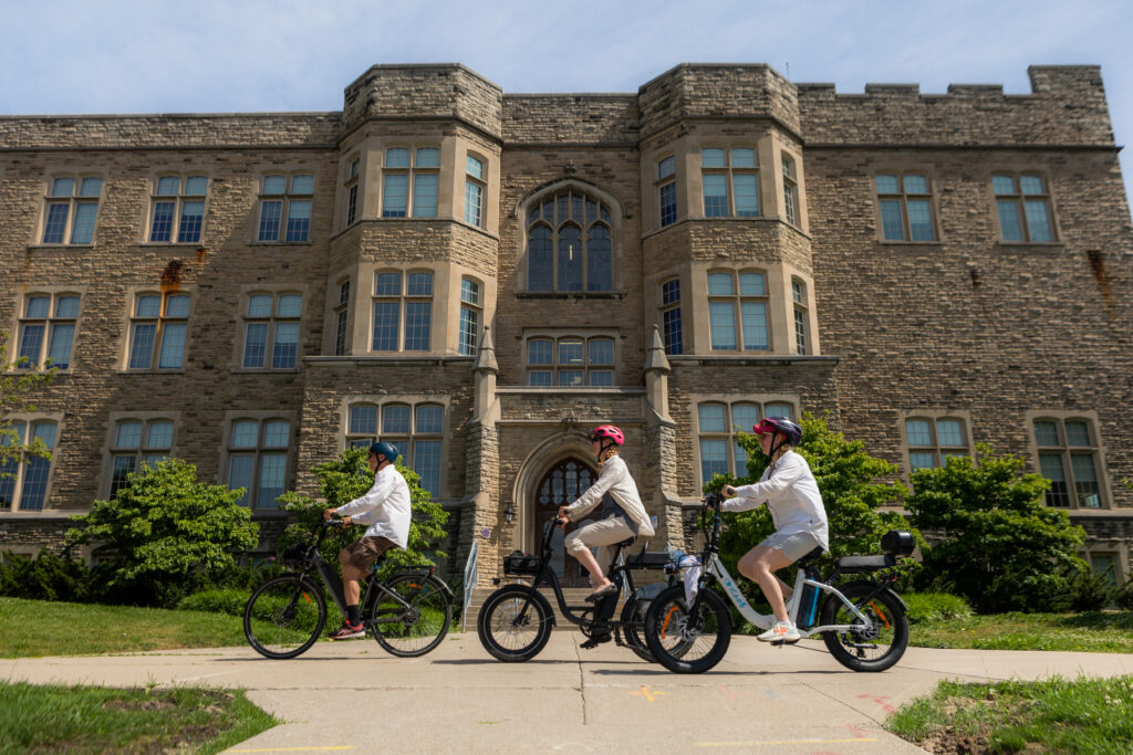 Three cyclists pass in front of a Gothic inspired building.