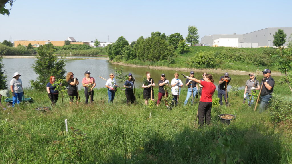 A group of tree planters receive instruction in a field, beside a river.