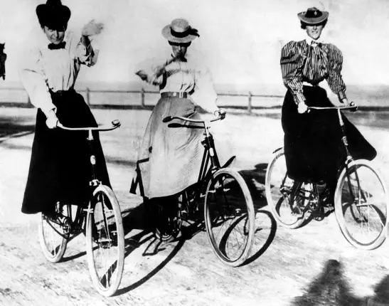 Women in dresses and hats riding bicycles, circa 1900. 