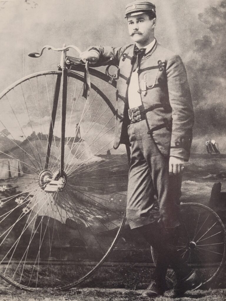 Man standing by a high-wheeler in 1880s.