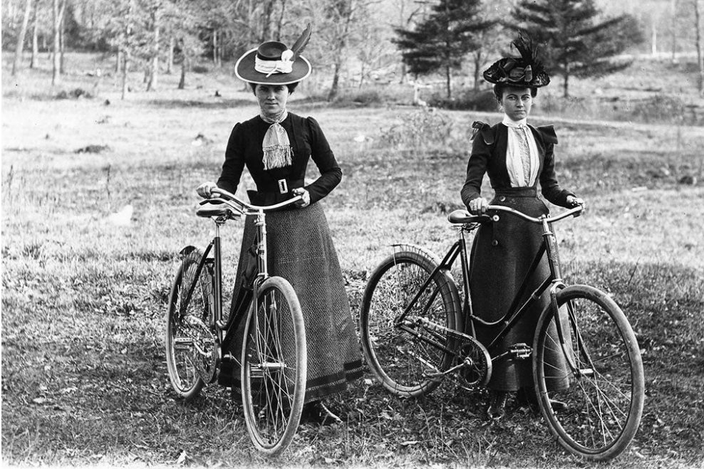 Women in dresses and hats, astride bicycles, circa 1900.