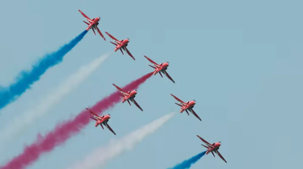 Seven red jets flying in formation, trailing red, white, and blue smoke for the London Airshow.