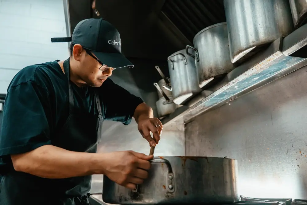 Male kitchen worker stirs the food in a large pot on a stove top.